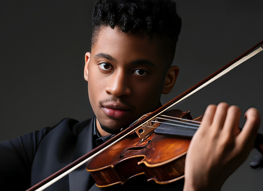 Violinist Irain Quirós, recipient of the 2025 Cecilia Concerts Developing Musician Award, poses with his violin against a dark background. He wears a black suit and holds the violin up near his shoulder while drawing the bow across the strings. His expression is calm and focused, and the image highlights both his face and the warm wood tones of the instrument.