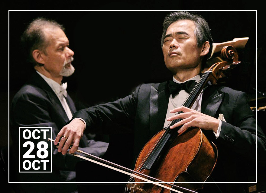 Two men are captured in a concert setting. The man on the left, with graying hair and a beard, wears a white dress shirt with a black bowtie and suit. He appears to be observing. On the right, a man with dark hair, in a black tuxedo with a white dress shirt and black bowtie, is deeply engrossed in playing a cello. He has his eyes closed, expressing intense emotion. The background is dark, emphasizing the performers. In the lower-left corner, there's a graphic depicting a calendar page showing "OCT 28".