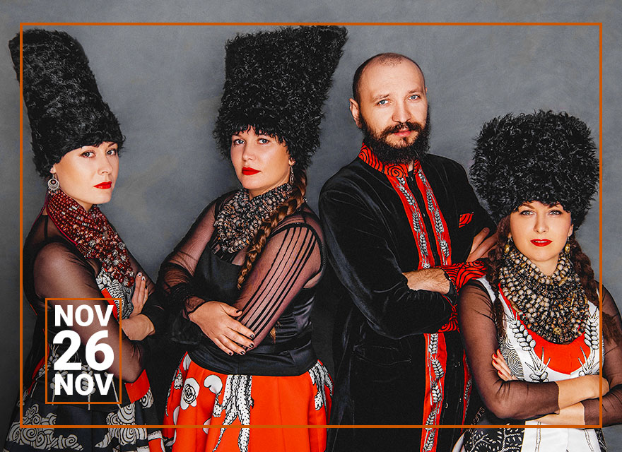 This image shows the music quartet DakhaBrakha, identifiable by their distinctive stage attire. Three women and one man stand side by side. The women are dressed in red and white garments with intricate patterns, accented with black scarves and unique, tall black fur hats. Their serious expressions and direct gazes give them a commanding presence. The man, also in black, sports a similar fur hat but in a lighter color, with a full beard and a contemplative expression. The group's traditional yet stylized outfits reflect their musical fusion of Ukrainian folk with global rhythms.