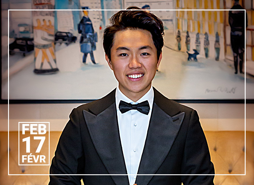 Violinist Hao Zhou stands confidently in a luxurious lobby, holding his violin. He is smartly dressed in a classic black tuxedo with a bow tie, radiating a cheerful smile. A comfortable-looking tufted sofa is behind him and a whimsical cityscape painting decorates the wall, contributing to the elegant atmosphere.