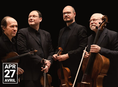 A photograph of Quatuor Élysée, a string quartet, featuring four musicians dressed in black formal attire holding string instruments. The background is completely black, emphasizing their figures. All four musicians are smiling, conveying a sense of camaraderie and warmth.