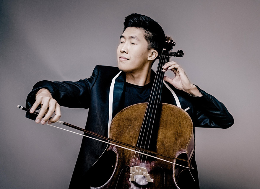 Cellist Bryan Cheng playing the cello with eyes closed, wearing a black jacket with white lapel detailing, against a neutral grey background.