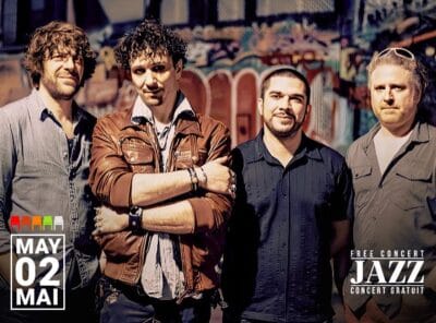 French jazz ensemble Beltuner poses in front of a vibrant, graffiti-covered wall. The four musicians stand close together, casually dressed, with relaxed expressions. A date graphic in the lower left corner reads "May 02 / Mai 02" alongside a row of colored seat icons indicating availability.