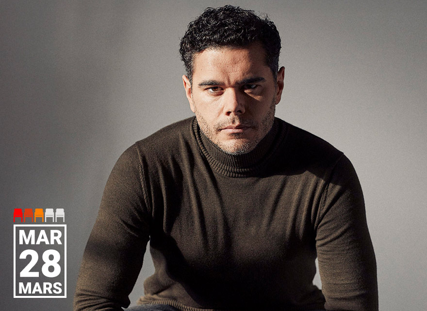 Baritone Elliot Madore is seated against a neutral background, wearing a dark olive-green turtleneck sweater. He gazes directly at the camera with a serious, intense expression. The lighting casts soft shadows, emphasizing the contours of his face and form. In the bottom left corner, white text reads "MAR 28 MARS."