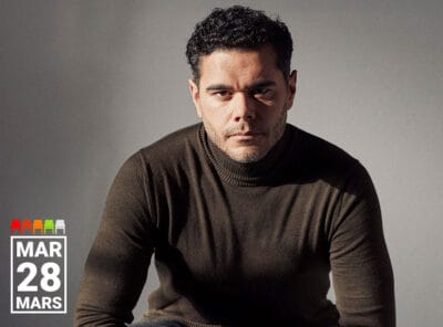 Baritone Elliot Madore is seated against a neutral background, wearing a dark olive-green turtleneck sweater. He gazes directly at the camera with a serious, intense expression. The lighting casts soft shadows, emphasizing the contours of his face and form. In the bottom left corner, white text reads "MAR 28 MARS."