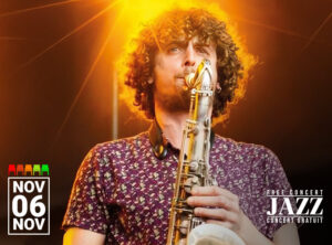 Musician Illyes Ferfera of the Illyes Ferfera Quartet performing on stage. He is playing a saxophone under a dramatic orange spotlight, with curly hair and wearing a patterned shirt. A date, November 6, is prominently displayed in the lower left corner, along with a row of colored seat icons indicating availability.