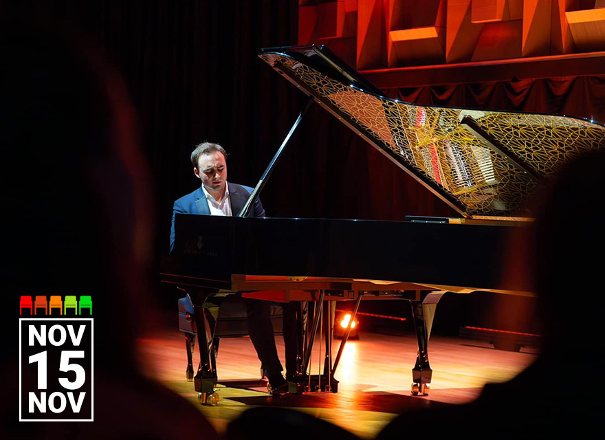 Pianist Illia Ovcharenko performs on a grand piano under warm stage lighting. The piano lid features an intricate decorative pattern that reflects the stage lights, adding a golden glow. He is dressed formally in a dark suit and white shirt, with an expressive, focused look. The audience is partially visible in shadowed silhouette in the foreground. In the bottom left corner, white text in a black box reads "NOV 15 NOV."