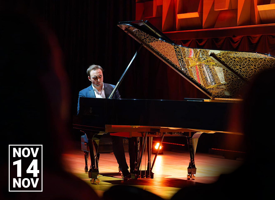 Ukrainian pianist Illia Ovcharenko performs on a grand piano under warm stage lighting, with the piano lid open to reveal its intricate gold lattice design. The image includes a date graphic reading “Nov 14” with coloured chair icons.