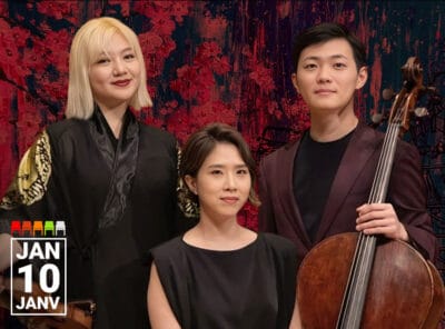 Three classical musicians pose indoors in a concert hall against an abstract red and black backdrop. On the left, a woman with shoulder-length blonde hair wears a black robe with gold designs on the sleeves and holds a violin. In the center, a seated woman with short dark hair wears a sleeveless black dress and looks calmly at the camera. On the right, a man with short black hair wears a dark plum suit and holds a cello upright beside him. In the bottom left corner, white text reads "JAN 10 JANV."
