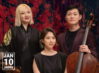 Three classical musicians pose indoors in a concert hall against an abstract red and black backdrop. On the left, a woman with shoulder-length blonde hair wears a black robe with gold designs on the sleeves and holds a violin. In the center, a seated woman with short dark hair wears a sleeveless black dress and looks calmly at the camera. On the right, a man with short black hair wears a dark plum suit and holds a cello upright beside him. In the bottom left corner, white text reads "JAN 10 JANV."
