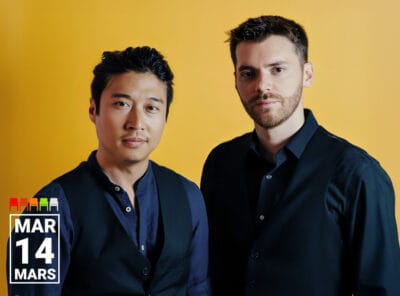 Two musicians stand side by side against a solid yellow background. On the left, a man with short black hair wears a dark navy shirt and black vest, looking directly at the camera with a slight smile. On the right, a man with short brown hair and a trimmed beard wears a black dress shirt and matching vest, gazing at the camera with a neutral expression. In the bottom left corner, white text reads "MAR 14 MARS."