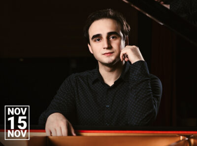 Pianist Illia Ovcharenko sits at a grand piano, resting one arm on the instrument and looking toward the camera with a calm, thoughtful expression. He wears a dark patterned shirt against a softly lit concert hall background. A small graphic in the lower left corner displays the date November 15.