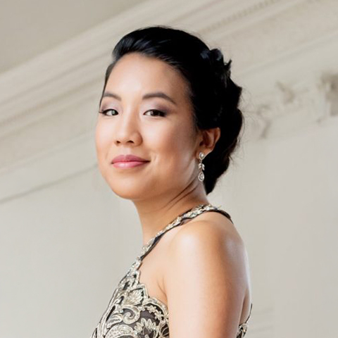 A portrait of pianist Silvie Cheng, shown from the waist up in an elegant, sleeveless patterned gown. She faces the camera with a calm and confident expression, her dark hair styled in a smooth updo.