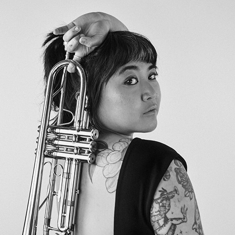 Black and white portrait of trumpeter Abbey Frias. She faces the camera over her shoulder while holding a trumpet across her back, raised behind her head with one hand. She has short dark hair with bangs, visible tattoos on her arm and shoulder, and wears a sleeveless black top.