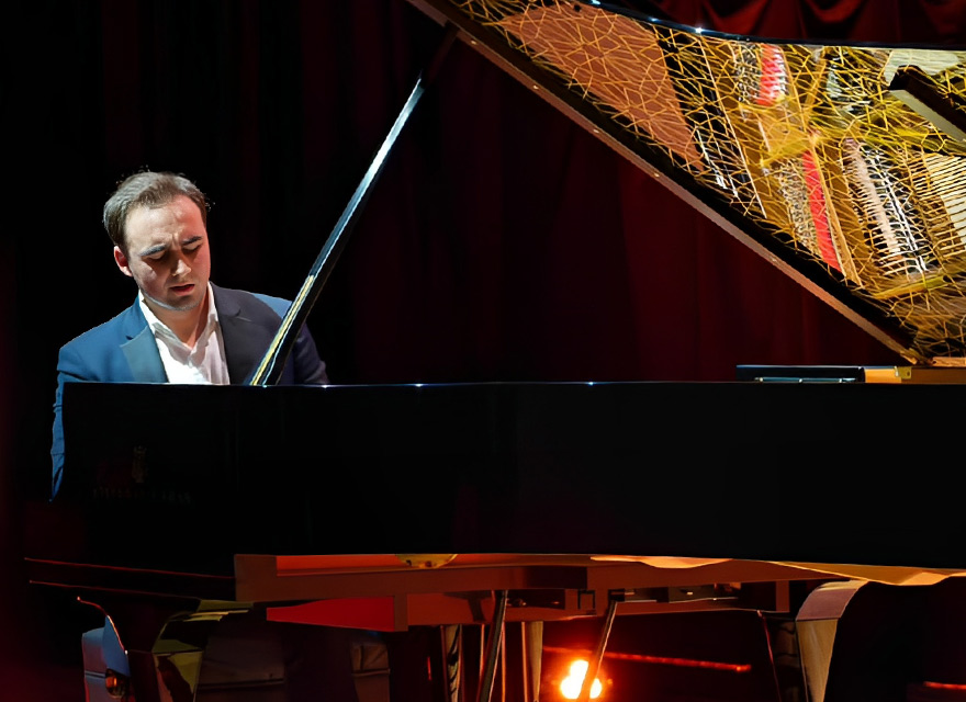 Pianist Illia Ovcharenko performs at a grand piano on stage, with the piano lid open and warm lighting against a dark red curtain backdrop.